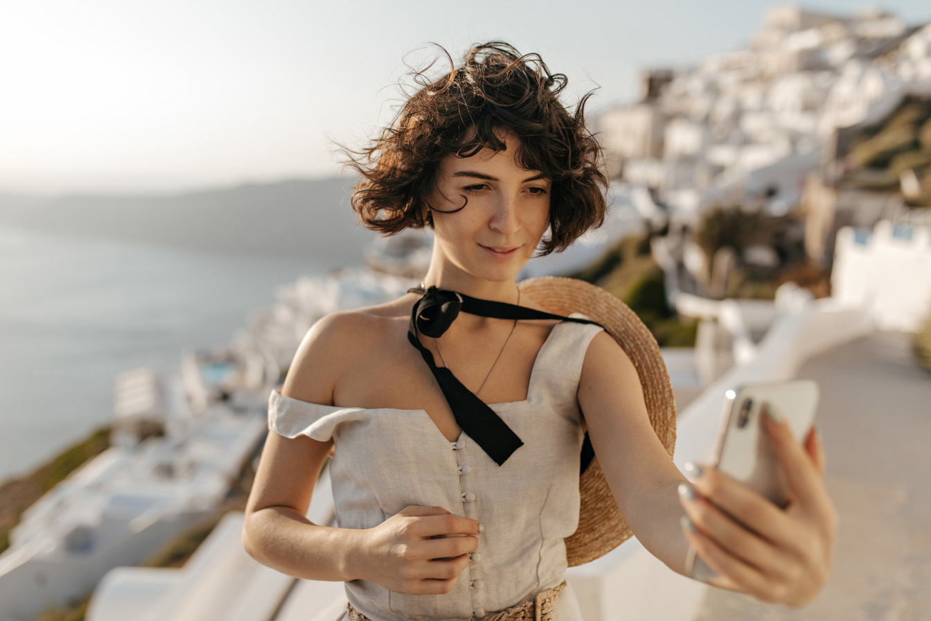 brunette-curly-woman-beige-dress-straw-hat-takes-selfie-sea-city-wall