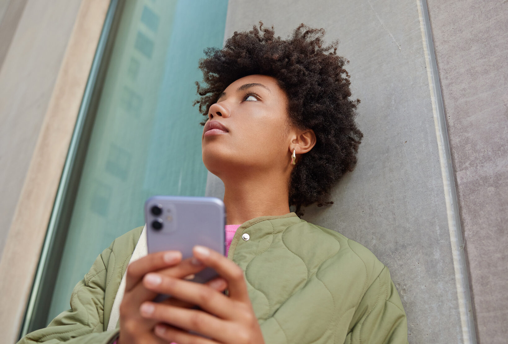 woman-uses-phone-chatting-with-fried-holds-mobile-phone-focused-somewhere-wears-casual-jacket-poses-near-wall-modern-city-building